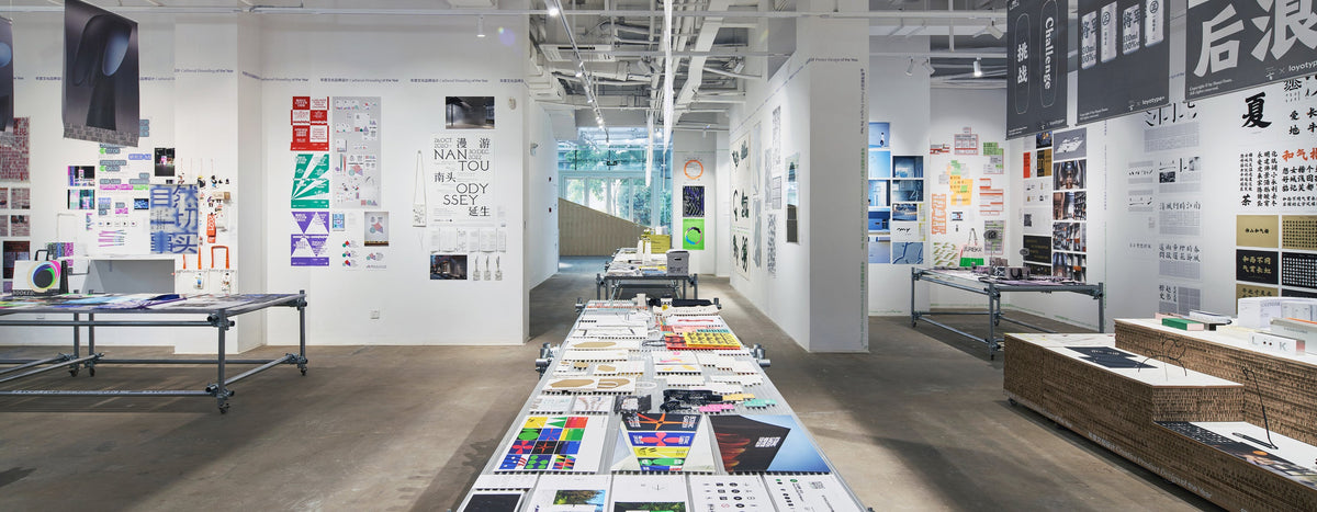 Wide view of an exhibition with printed designs on long tables and gallery walls.