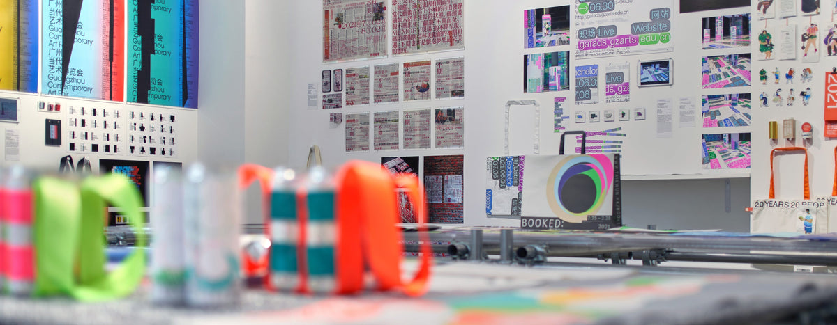 Colourful design objects on a gallery table with printed artwork on the wall behind.